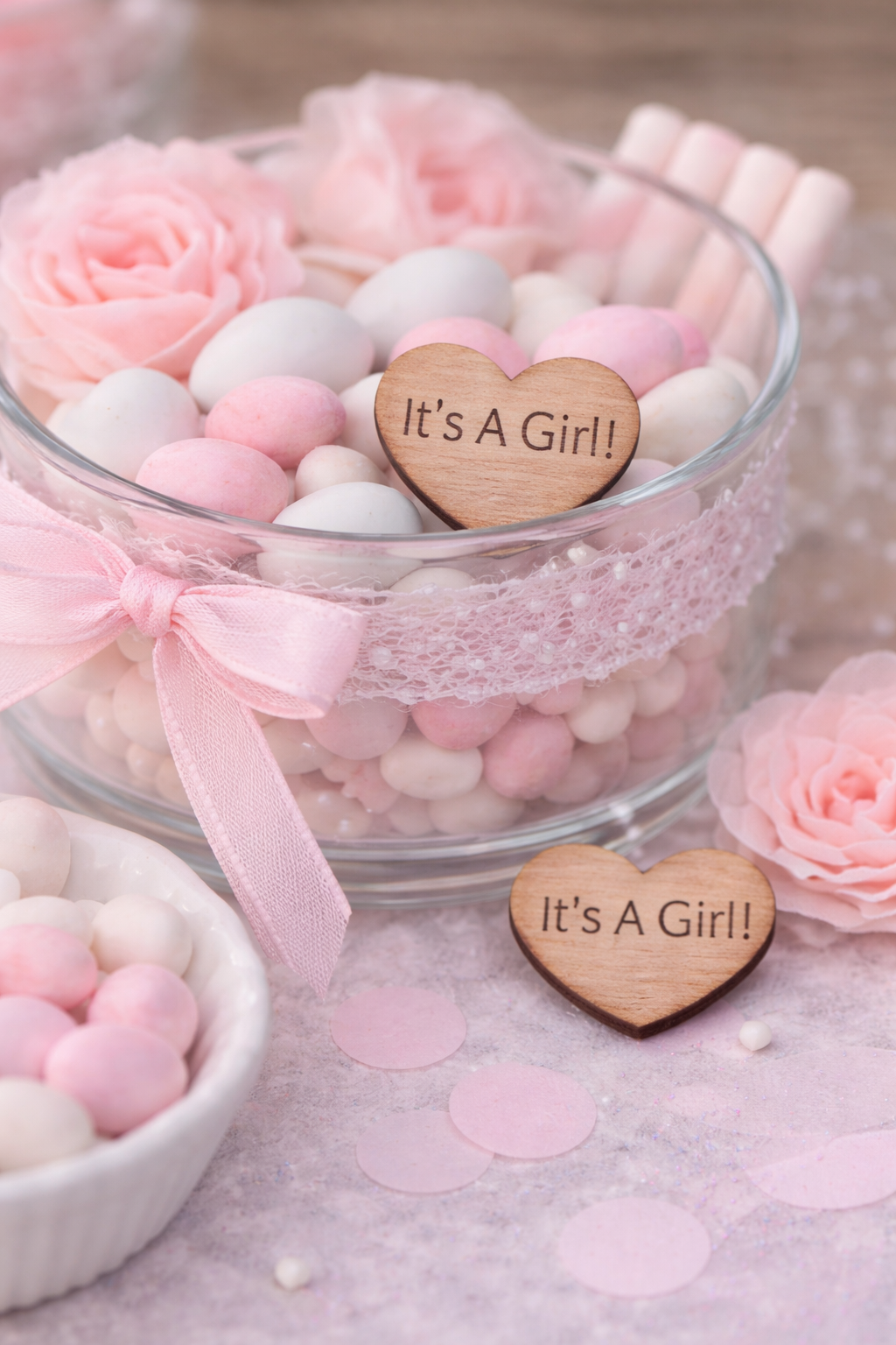 Decorative glass bowl with pink and white candies and 'It's a Girl!' heart signs, surrounded by pink flowers.