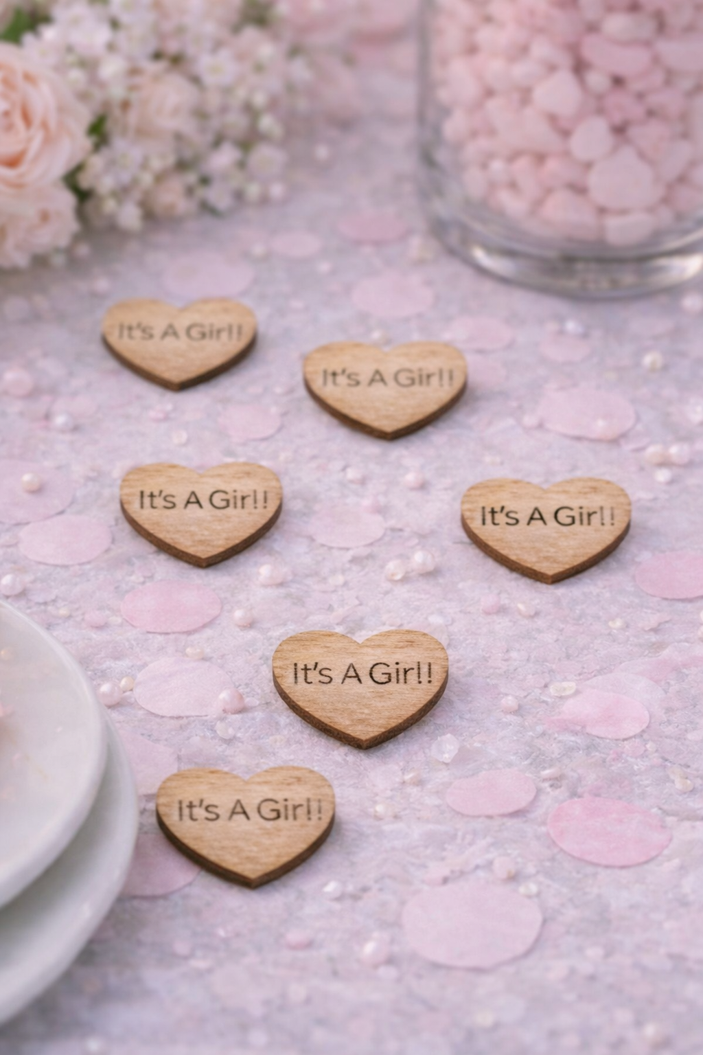 Wooden heart-shaped decorations with 'It's a Girl!' text on a light surface with flowers and pink hearts.