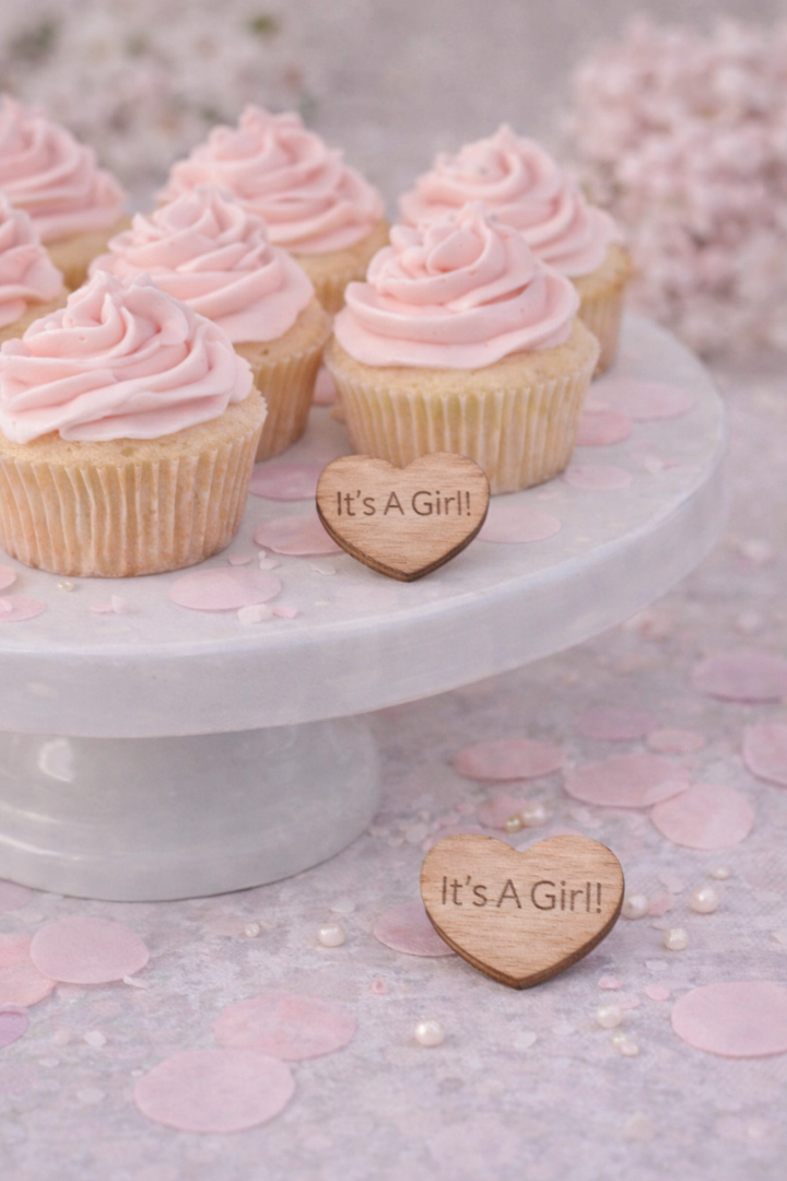 Cupcakes with pink frosting on a cake stand, surrounded by 'It's a Girl!' heart signs.